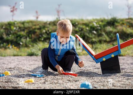 Un enfant blond mignon joue avec des jouets de sable près de l'oscillation d'équilibre dans la cour. Surface sûre avec de petits cailloux dans l'aire de jeux. Banque D'Images