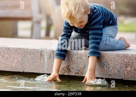 Joyeux garçon touchant l'eau dans la fontaine. Un garçon se réjouit en été dans une belle fontaine. Des loisirs d'été actifs pour les enfants en ville. Banque D'Images