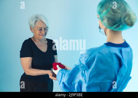 Femme médecin en uniforme médical, masque et capuchon médical donnant un coeur humain artificiel à une vieille dame aux cheveux gris portant un ensemble et des lunettes noirs. Prise de vue en studio. Photo de haute qualité Banque D'Images