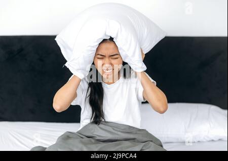 Une fille asiatique irritée, assise dans un lit à la maison, couvre ses oreilles avec un oreiller, tente de dormir, se ferme du bruit, a un mauvais rêve, rêve agité. Concept sommeil et repos Banque D'Images