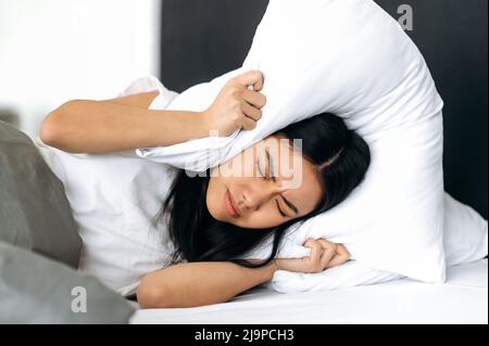 Concept sommeil et repos. Triste fille chinoise, couchée dans un lit à la maison, couvre ses oreilles avec un oreiller, tente de dormir, se ferme du bruit, a un mauvais rêve, rêve agité Banque D'Images