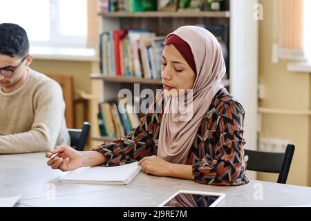 Portrait d'une femme musulmane moderne portant le hijab assise à table dans la bibliothèque et prenant des notes pendant la leçon pour les immigrants Banque D'Images