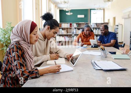 Groupe d'étudiants ethniquement divers assis à table regardant des vidéos éducatives sur une tablette numérique et faisant des tâches d'écriture Banque D'Images
