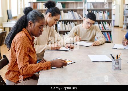 Groupe d'élèves immigrants ethniquement divers qui apprennent l'anglais assis dans une bibliothèque faisant un test de grammaire Banque D'Images