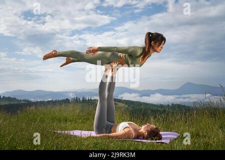 Vue de deux femmes faisant ensemble du yoga à l'extérieur, se tenant les unes les autres. Femme allongé sur un tapis de yoga et gardant l'équilibre par les jambes avec sa petite amie. Carpathian Mountains en arrière-plan. Banque D'Images