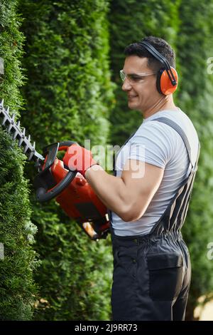 Vue latérale d'un homme caucasien heureux tailleur haie avec coupe électrique pendant la journée ensoleillée. Jardinier compétent portant des lunettes de sécurité, des écouteurs et des gants. Banque D'Images