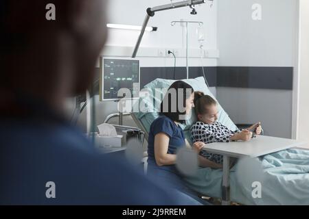 Mère assise à côté du lit du patient dans la salle de clinique pédiatrique pendant que la petite fille joue sur un smartphone. Jeune patient joyeux appréciant le divertissement du téléphone mobile tout en étant assis dans le service de récupération clinique. Banque D'Images