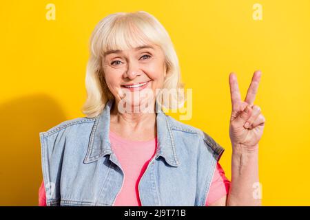 Photo de drôle douce dame pensioner vêtue gilet de denim montrant V-sing isolé couleur jaune fond Banque D'Images