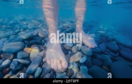 Pieds de femmes debout sur un fond de cailleboise dans l'eau de mer. Photo sous l'eau. Banque D'Images