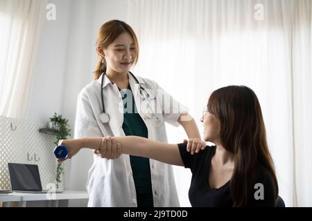 Jeune femme patiente utilisant des haltères avec des bras étirés tout en obtenant le traitement de physiothérapeute. Banque D'Images