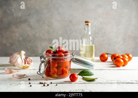 Tomates marinées dans un pot en verre avec de l'ail, de l'huile, des tomates fraîches, du poivre et des feuilles de Laurier sur une table en bois blanc à fond gris Banque D'Images
