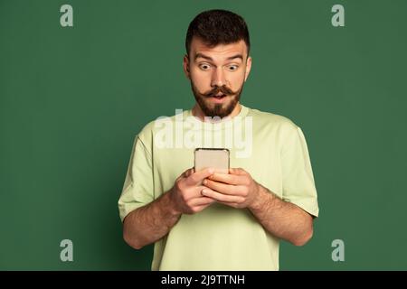 Portrait d'un jeune homme émotif regardant le téléphone avec une expression choquée isolé sur fond vert de studio. Message expressif Banque D'Images