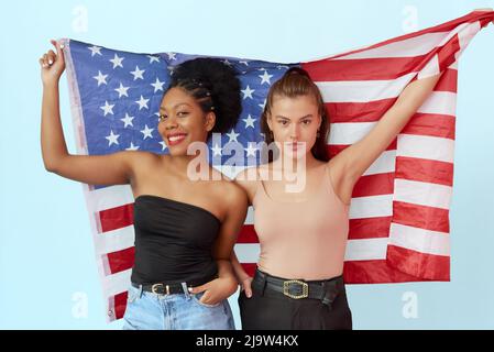 Femme afro-américaine et femme caucasienne portant le drapeau américain en studio sur fond bleu. Le concept de l'unité des différentes nations et indépendants Banque D'Images