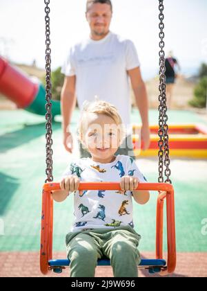 Un petit garçon blond caucasien de 3 ans se balance sur une balançoire lors d'une chaude journée d'été ensoleillée dans l'aire de jeux par son jeune père. Banque D'Images