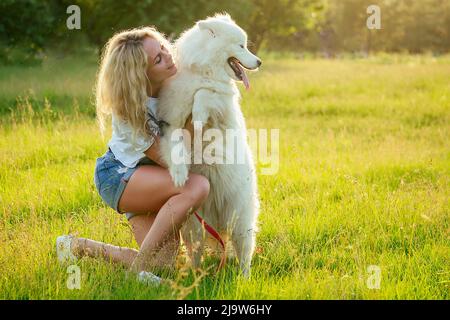 belle et charmante femme tortueuse blonde souriante en combinaison en denim sont assis à un baiser de verre un chien doux blanc de samoyed dans le parc d'été Banque D'Images