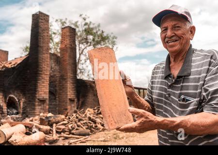 Homme âgé dans un chapeau souriant et regardant la caméra tenant une tuile d'argile dans ses mains devant un four de brique au Nicaragua Banque D'Images