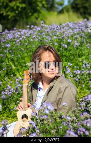 Portrait spontané d'une jeune femme souriante heureuse avec ukulele profitant de la lumière du soleil dans la prairie à fleurs au printemps Banque D'Images