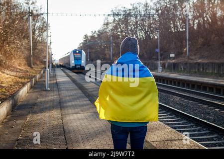 Femme enveloppée dans le drapeau ukrainien debout à la plate-forme de la gare et regardant arriver le train. Concept de crise humanitaire et de sortie de refuge Banque D'Images