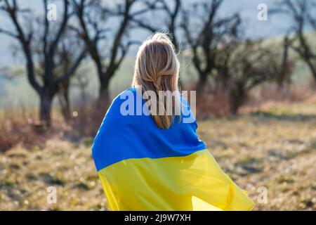 Femme couverte par un drapeau ukrainien debout sur le terrain. Concept d'espoir et de liberté pour le pays d'Ukraine Banque D'Images