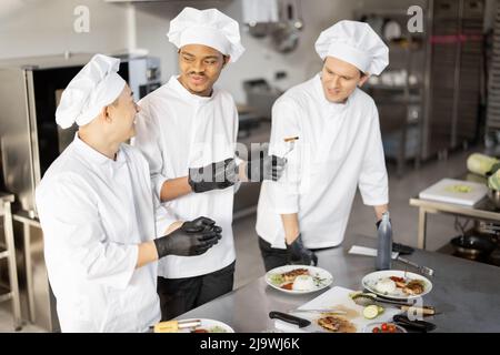 Trois cuisiniers avec différentes spécialités goûtant des plats cuisinés, inventant de nouveaux plats pour un menu. Chef asiatique avec des cuisiniers latino et européens en uniforme travaillant ensemble. Concept de travail d'équipe et cuisine haute Banque D'Images