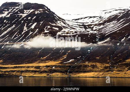 Les montagnes enneigées des fjords de l'Est de l'Islande portent plusieurs cascades ; une petite maison se dresse sur la rive en contrebas. Banque D'Images