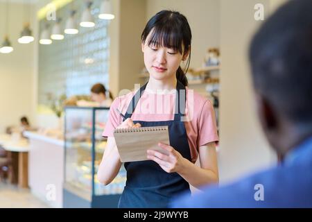 Jeune serveuse asiatique en tablier prenant des notes dans le bloc-notes tout en prenant la commande de l'homme au restaurant Banque D'Images