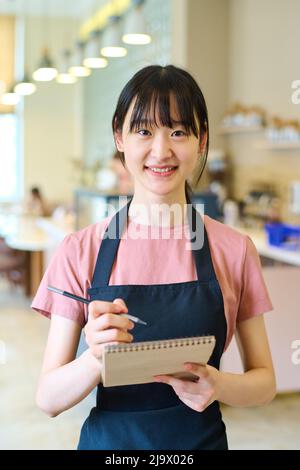 Portrait de la jeune serveuse asiatique en tablier souriant à l'appareil photo tout en prenant la commande au restaurant Banque D'Images