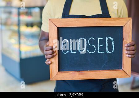 Gros plan du serveur afro-américain en uniforme tenant le tableau avec panneau fermé tout en se tenant dans le café Banque D'Images