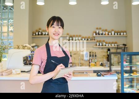 Portrait de la jeune serveuse asiatique en tablier souriant à l'appareil photo et prenant des notes dans le bloc-notes tout en se tenant dans le café Banque D'Images