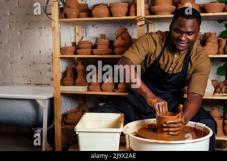 course mixte afro mâle potter avec tablier noir et chemise sombre élégante assis à la table d'atelier potter's wheel , application de glaçure sur l'argile fait à la main marron Banque D'Images