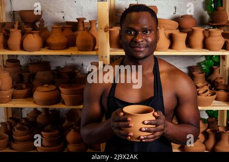 course mixte afro mâle potter avec tablier noir et chemise sombre élégante assis à la table d'atelier potter's wheel , application de glaçure sur l'argile fait à la main marron Banque D'Images