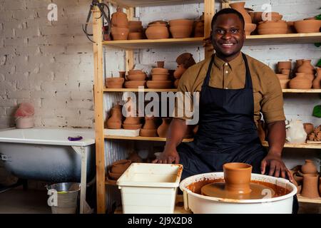 course mixte afro mâle potter avec tablier noir et chemise sombre élégante assis à la table d'atelier potter's wheel , application de glaçure sur l'argile fait à la main marron Banque D'Images