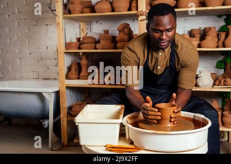 course mixte afro mâle potter avec tablier noir et chemise sombre élégante assis à la table d'atelier potter's wheel , application de glaçure sur l'argile fait à la main marron Banque D'Images