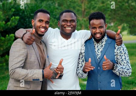 un groupe de trois hommes noirs dans l'élégance convient à une réunion dans un parc d'été. Les amis afro-américains le salut d'un homme d'affaires hispanique pose le travail d'équipe à l'extérieur Banque D'Images