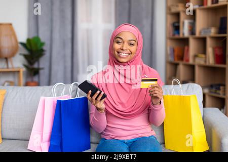 Jolie femme musulmane noire avec sacs cabas, carte de crédit et smartphone shopping en ligne de la maison, assis sur un canapé Banque D'Images