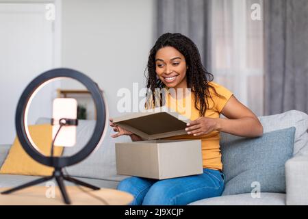 Bonne femme noire de blogueur de diffusion tout en ouvrant la boîte de livraison, assis sur le canapé devant le smartphone et la bague de lampe Banque D'Images