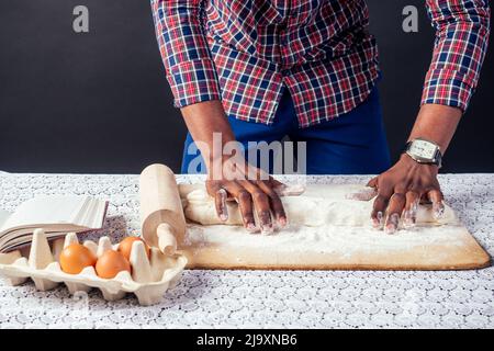 Afro-américain cuisinier chef se vante les mains de pâtisserie dans la farine pâtisserie .les mains de l'homme pâte roulante punaise gâteau, oeufs et livre de recettes sur la table Banque D'Images