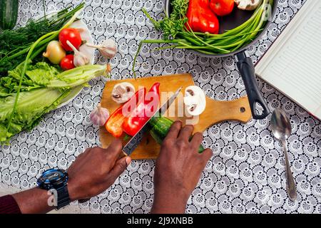 gros plan des mains de l'homme coupes afro-américaines légumes friture salade poivre, champignons, tomate en cuisine livre de recettes sur la table .vegan nourriture saine Banque D'Images