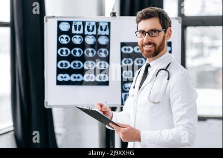 Fier homme expérimenté de médecine générale caucasienne médecin, chirurgien, pulmonologieist, en uniforme médical, avec des lunettes et un stéthoscope sur les épaules, se tenir dans la clinique, regardant la caméra, souriant Banque D'Images