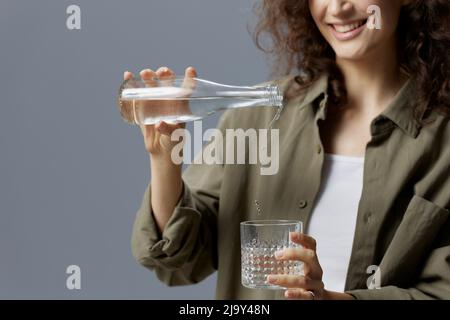 Joyeux sourire curly belle femme dans décontracté kaki vert chemise verse l'eau de la bouteille dans le verre posant isolé sur fond gris bleu. En bonne santé Banque D'Images