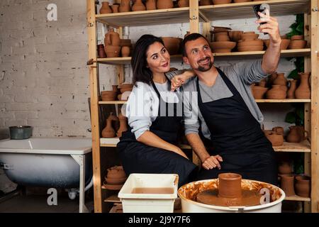 portrait d'une belle femme de potier et homme heureux faisant le selfie au téléphone. famille d'artistes travaillant avec l'argile sur une roue de potier dans la poterie Banque D'Images