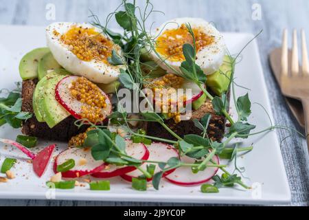 Pain grillé, œufs durs, tranche d'avocat, petits pois et radis frais sur une assiette blanche dans un café, gros plan, heure du petit déjeuner Banque D'Images