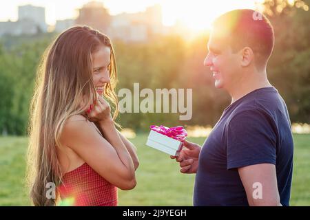 amant homme donne une boîte cadeau à embarrassée belle blonde long cheveux femme dans l'amour en soirée cocktail robe dans le parc d'été avec l'herbe Banque D'Images