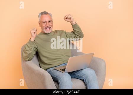 Photo de cool Elder gris coiffure homme assis avec ordinateur portable porter des lunettes pull jeans isolés sur fond beige Banque D'Images