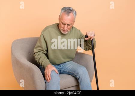 Photo de l'homme de cheveux gris plus âgé stressé s'asseoir avec le bâton porter pull jeans isolés sur fond beige Banque D'Images