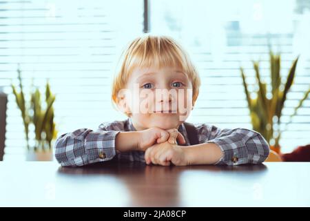 Un garçon de 3 ans avec des cheveux blonds en chemise sourit. Un enfant heureux sur la table. Banque D'Images