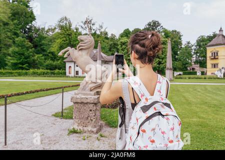 Une jeune fille méconnaissable à l'arrière prend des photos sur smartphone de monuments historiques, sculpture en pierre en forme de licorne, dans le parc Hellbrunn. Autriche, Banque D'Images