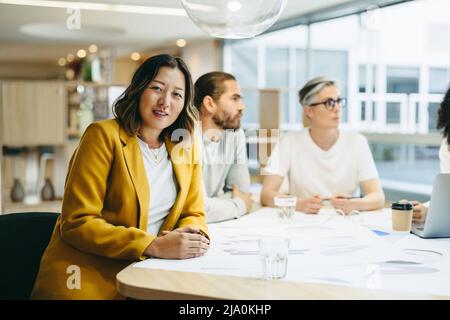 Une femme d'affaires gaie regardant la caméra lors d'une réunion avec ses collègues. Groupe de professionnels du design innovant partageant leur créativité Banque D'Images