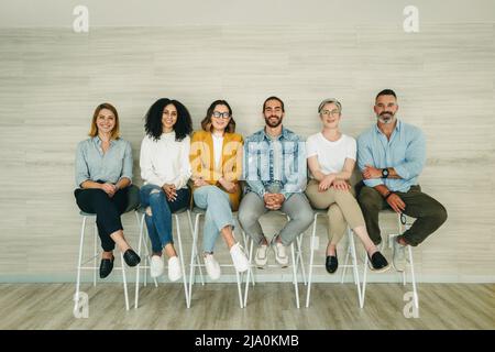 Les demandeurs d'emploi souriants sourient à la caméra tout en étant assis en file d'attente pour une entrevue. Groupe d'hommes d'affaires multiculturels en attente d'une occasion Banque D'Images