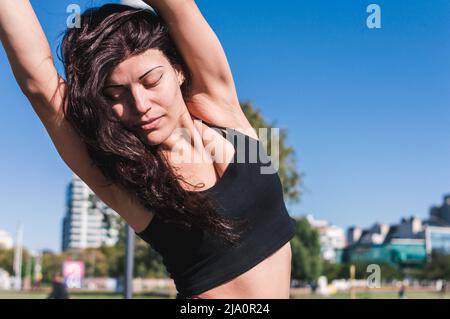 belle jeune femme latine caucasienne avec des yeux fermés à l'extérieur, étire ses bras et les épaules pour l'exercice, porte des vêtements de sport noirs, espace de copie de ciel Banque D'Images